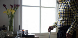 Elderly man standing with a cane next to a table with flowers and medication