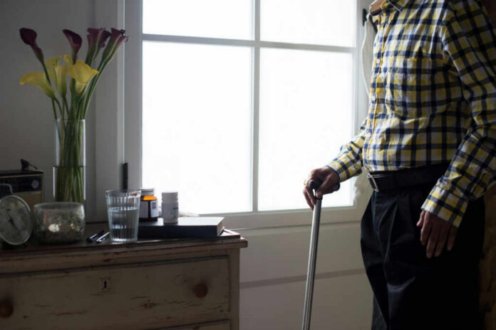 Elderly man standing with a cane next to a table with flowers and medication