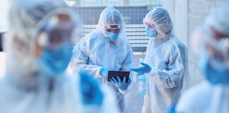 Healthcare workers in protective gear discussing information on a tablet outdoors
