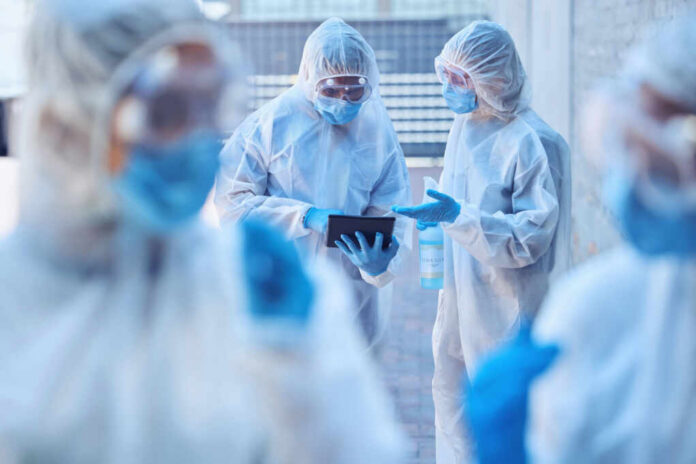 shutterstock2506350089jpg Healthcare workers in protective gear discussing information on a tablet outdoors