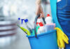 A person holding a blue bucket filled with cleaning supplies including spray bottles and cloths