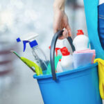 A person holding a blue bucket filled with cleaning supplies including spray bottles and cloths