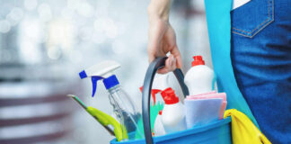 A person holding a blue bucket filled with cleaning supplies including spray bottles and cloths