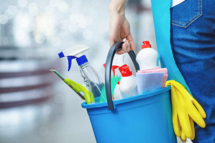 A person holding a blue bucket filled with cleaning supplies including spray bottles and cloths