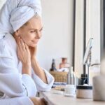 Sleep Mistakes Aging Skin Faster Than Sun Woman in a bathrobe with a towel on her head, smiling at her reflection in a mirror