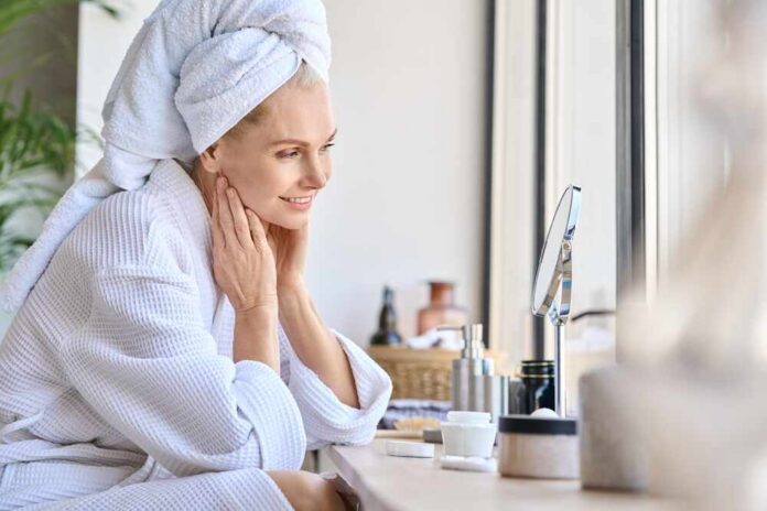Woman in a bathrobe with a towel on her head, smiling at her reflection in a mirror