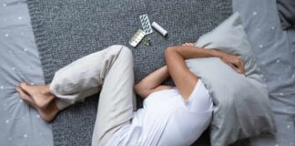 Person lying on a bed with a pillow over their head, surrounded by medication