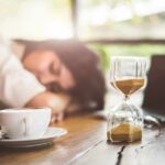 Hourglass on table with coffee cup and sleeping person in background