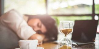 Hourglass on table with coffee cup and sleeping person in background
