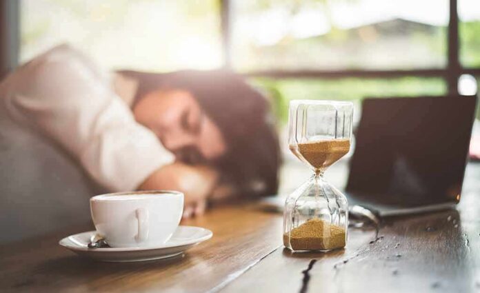 shutterstock_1145958767 Hourglass on table with coffee cup and sleeping person in background