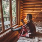 Woman wrapped in a blanket sitting on a bed by a cabin window