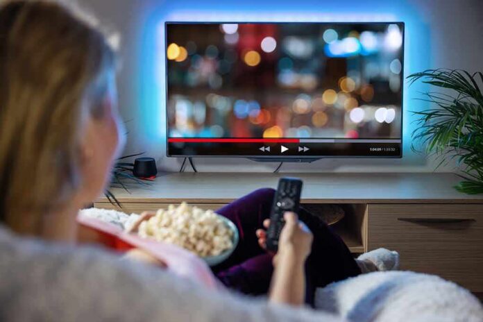shutterstock_1966720738-1 Person sitting on a couch watching a movie on a TV with popcorn in hand