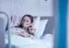 Child in hospital bed with oxygen mask holding teddy bear