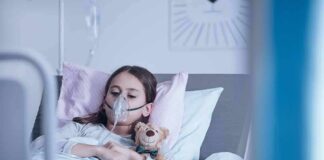 Child in hospital bed with oxygen mask holding teddy bear