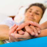 Hospitals Quietly Rewrite Dementia Playbook Caregiver holding hands with an elderly patient in bed