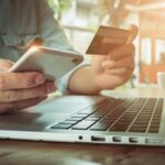Hands using smartphone and credit card beside a laptop