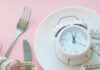 One Meal A Day: Benefits, Risks, and How it Works Pink alarm clock on a white plate with fork, knife, and measuring tape